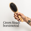 Hand med tatuering håller en Green Heads svinborstborste mot vit bakgrund – symbol för borstmetoden med 100 borsttag om dagen för en friskare hårbotten.